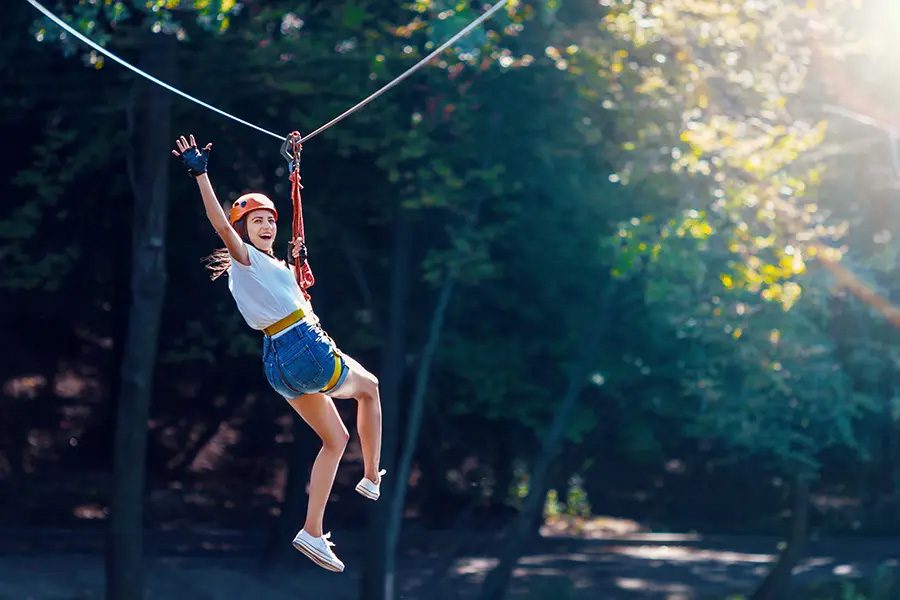 Adventure and Entertainment Insurance Cheerful Woman Waves as She is Gliding Along a Zipline in an Adventure Park